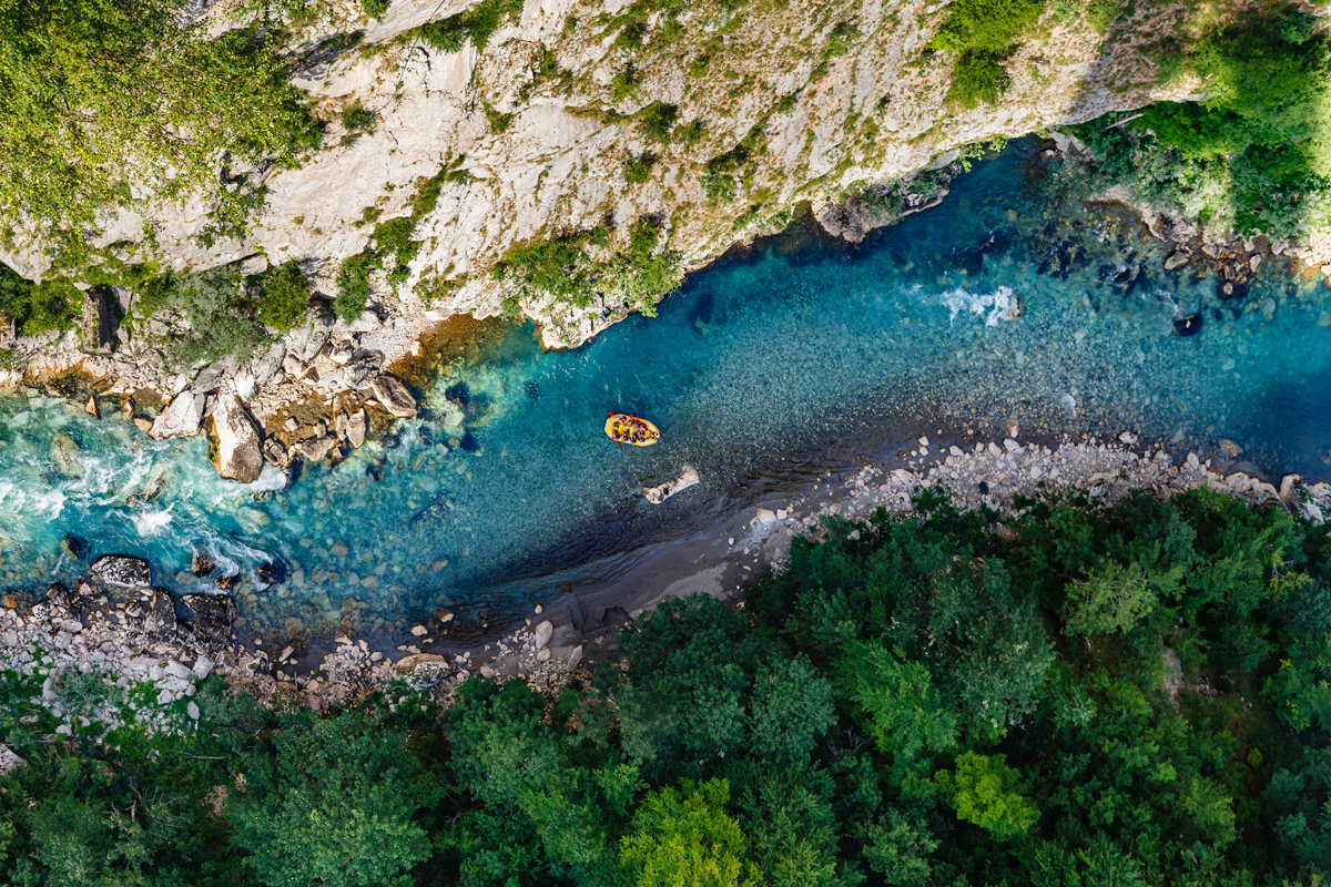 Aerial view of the blue river and a small yellow raft surrounded by sheer cliffs and lush forest in Tara River Canyon. 