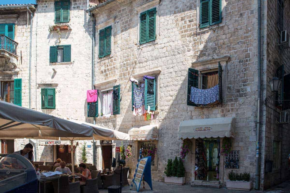 Washing hangs from windows with green shutters in Kotor Old Town Montenegro. There is a restaurant with umbrellas below the windows. 