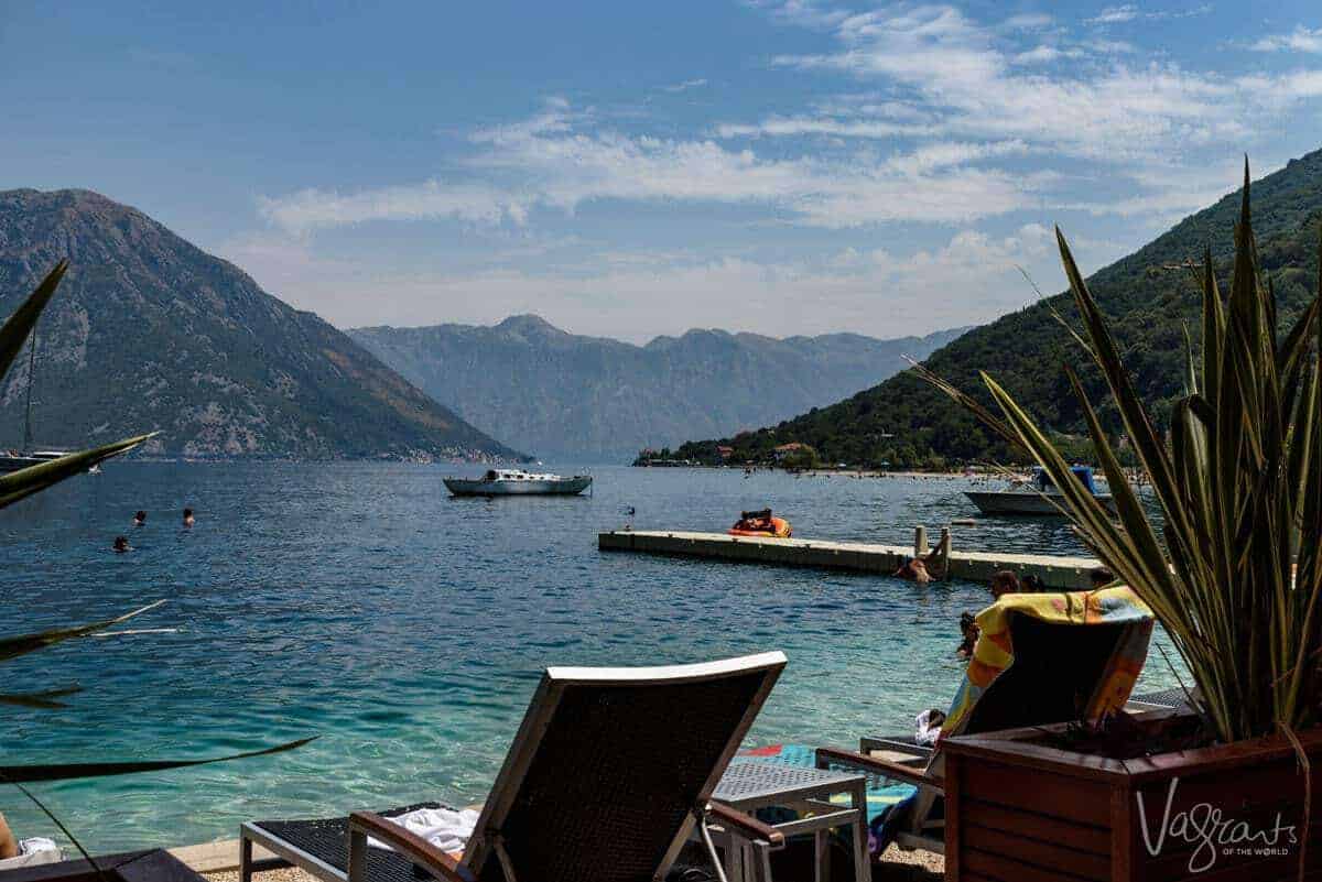Beach chairs on the edge of the clear water in Kotor Bay surrounded by mountains. People swim in the water and a boat is moored. 